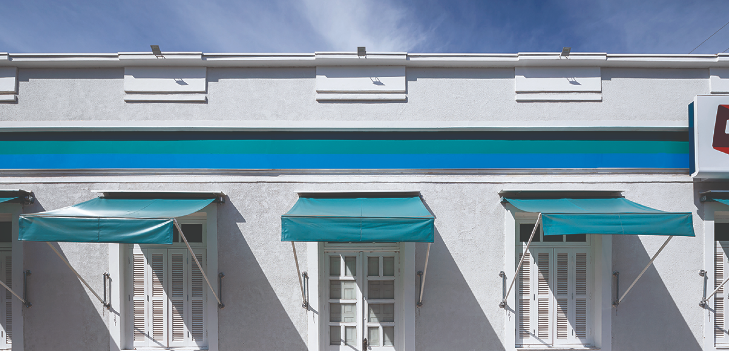 Fachada lateral da farmácia Drogaraia, com platibanda decorativa com detalhes em gesso branco e textura de cor gelo. O roda teto exibe faixas horizontais verde e azul entalhadas, acima de toldos verdes fixados por hastes de metal branco nas paredes. Abaixo é possível ver grandes janelas de madeira pintadas de branco, com venezianas e vidros retangulares. Ao fundo, o céu azul completa a cena.